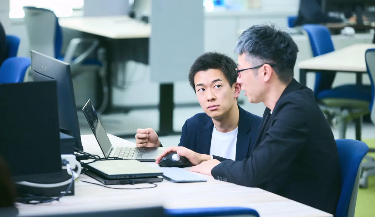 J.K. さんがチームメンバーのデスクにPCを持っていき、画面を見せながら意見を聞いている写真。