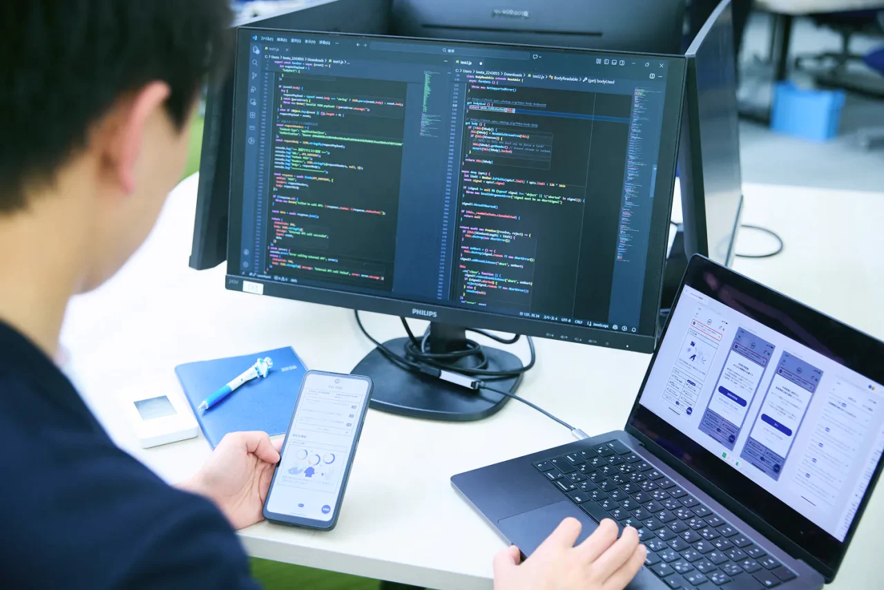 J.K. さんがスマートフォンを片手に、PCでプログラミングした結果を検証している写真。