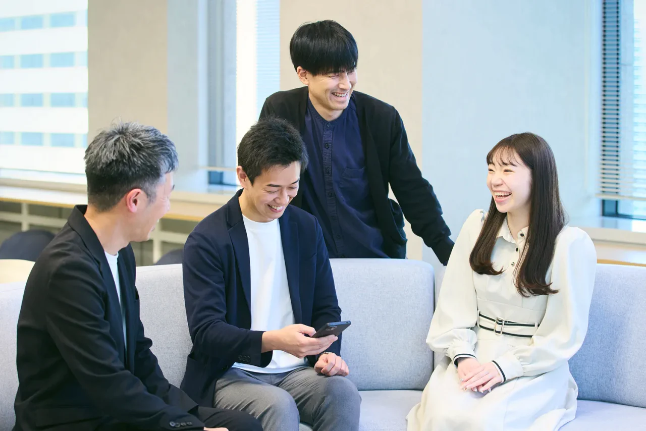 J.K.さんがメンバーの女性1人・男性2人と打ち合わせスペースでスマートフォンに映った開発画面を確認している写真。