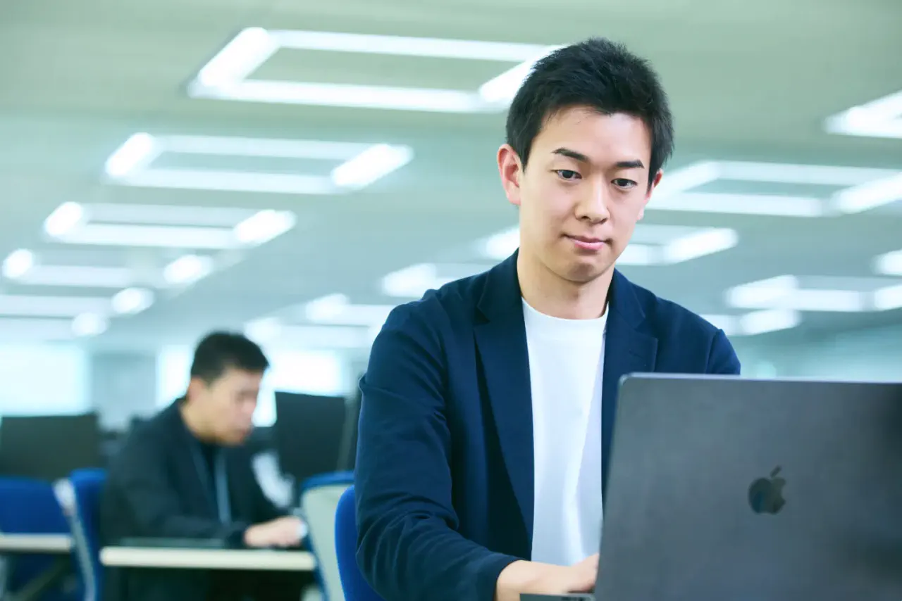 J.K. さんがデスクでPCを操作している写真。