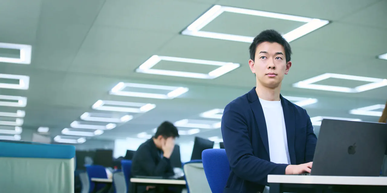 デスクに座りPCを操作しながら力強い視線で前を向く J.K. さんの写真