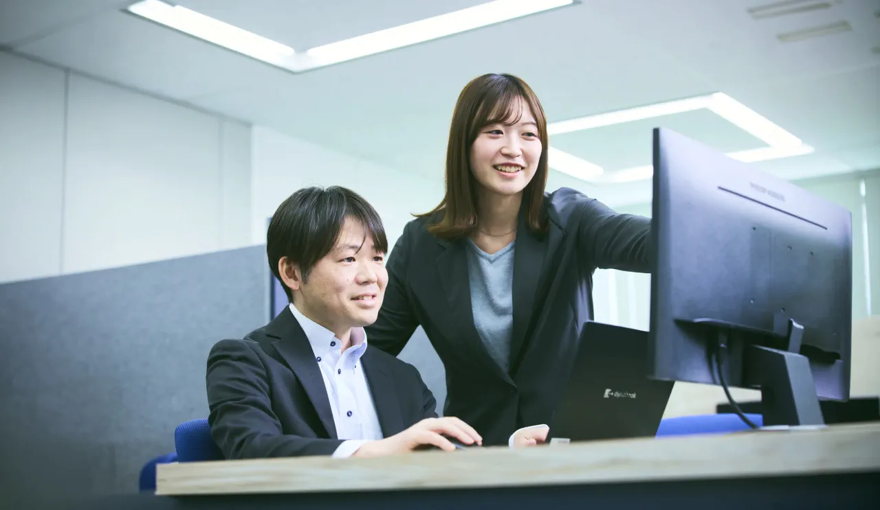 デスクでPCを作業をしているK.F.さんに女性社員1人がモニターを指さして会話している写真。