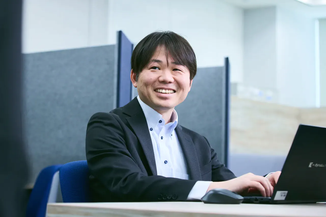 K.F. さんがデスクでPCで仕事をしていたところ、チームメンバーに話しかけられて笑顔で応対している写真。