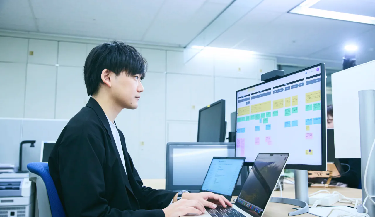 デスクでノートパソコンと外部モニターを接続して資料を作成している N.K.さんの写真。