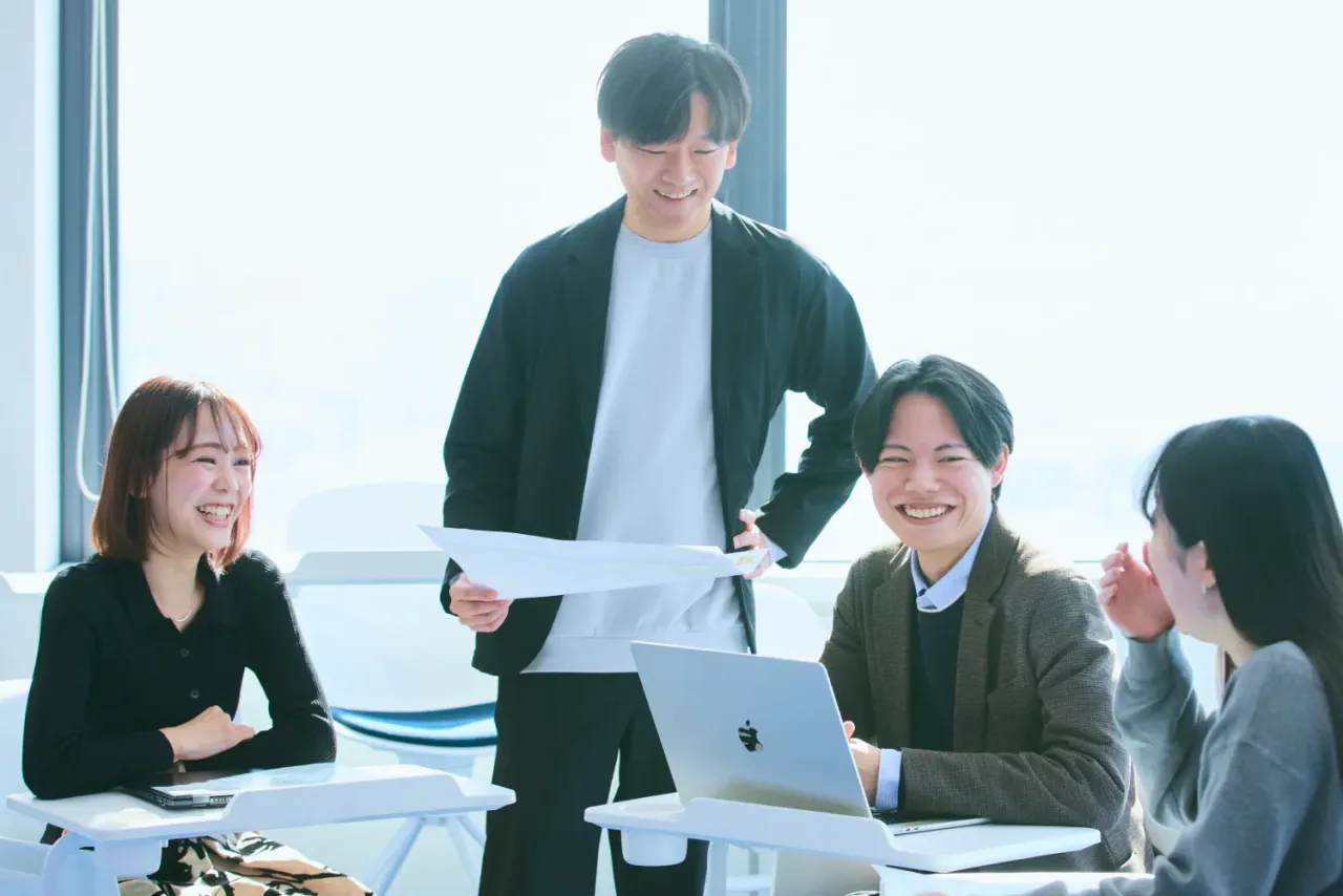 椅子に座っている男性1人、女性2人の間に立って資料を片手に打ち合わせをしている N.K.さんの写真。