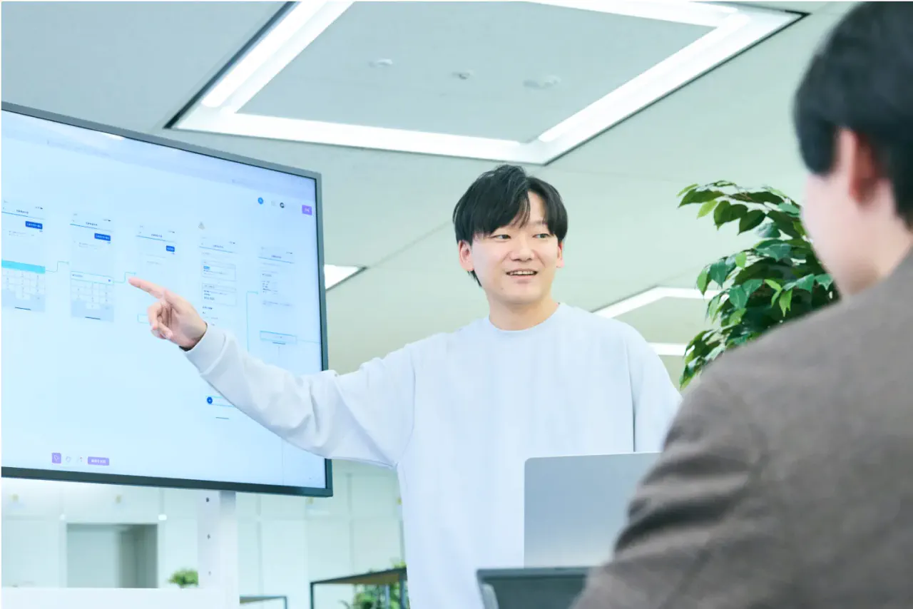 大型モニターに投影した資料を指さしながら社内メンバーと打ち合わせをしている N.K.さんの写真。