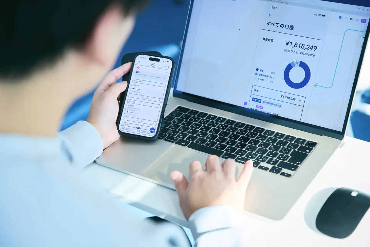 N.K.さんがスマートフォンを片手に、PCとあわせてそれぞれの画面でアプリデザインを確認している写真。