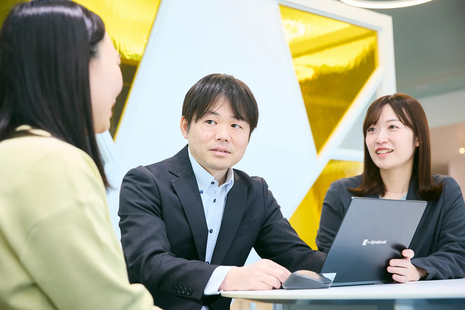 打ち合わせスペースで女性社員1人が発言している内容を、男性社員と女性社員それぞれ1名が聞いている様子の写真