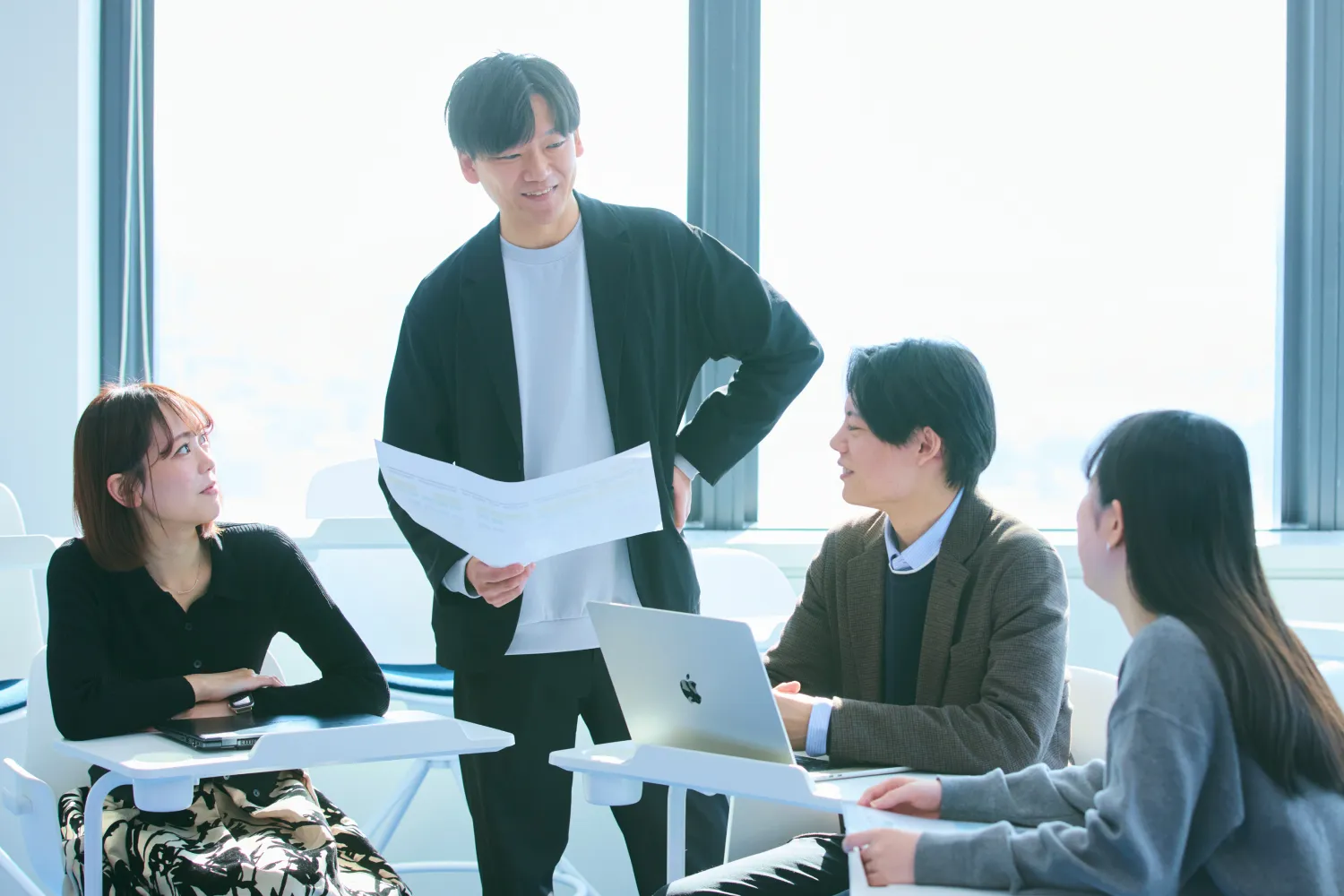 オフィスで資料を手に持った男性社員が座っている3名の同僚とパソコンを囲みながら談笑する様子の写真