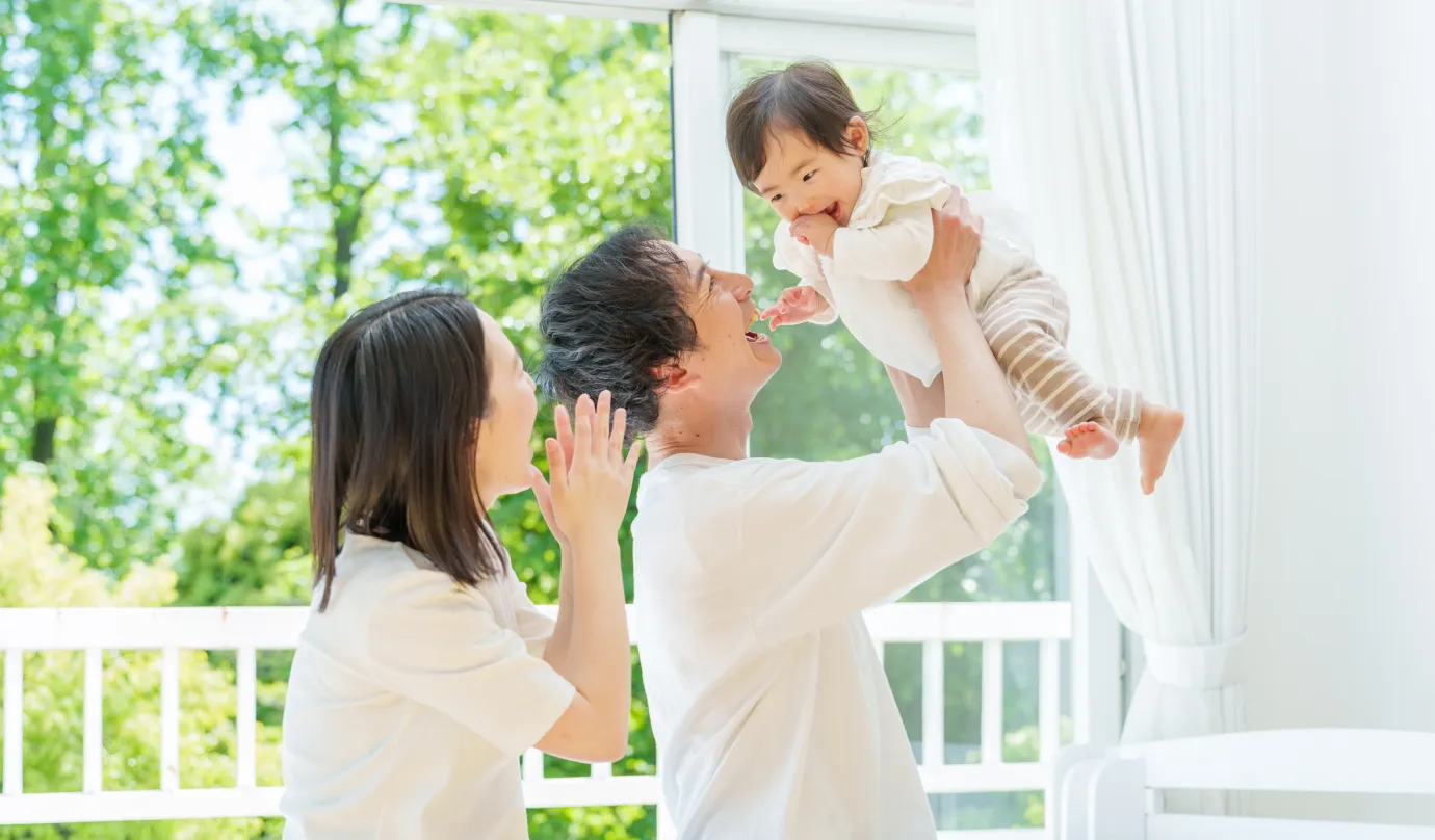 赤ちゃんを抱き上げて笑う父親と、それを見守る母親の家族の様子の写真