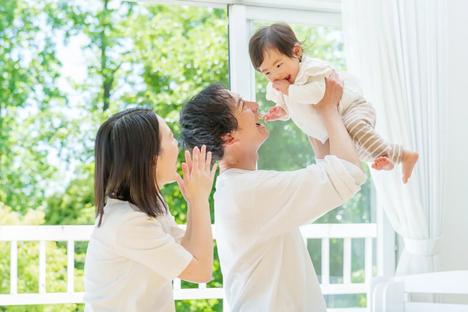 赤ちゃんを抱き上げて笑う父親と、それを見守る母親の家族の様子の写真
