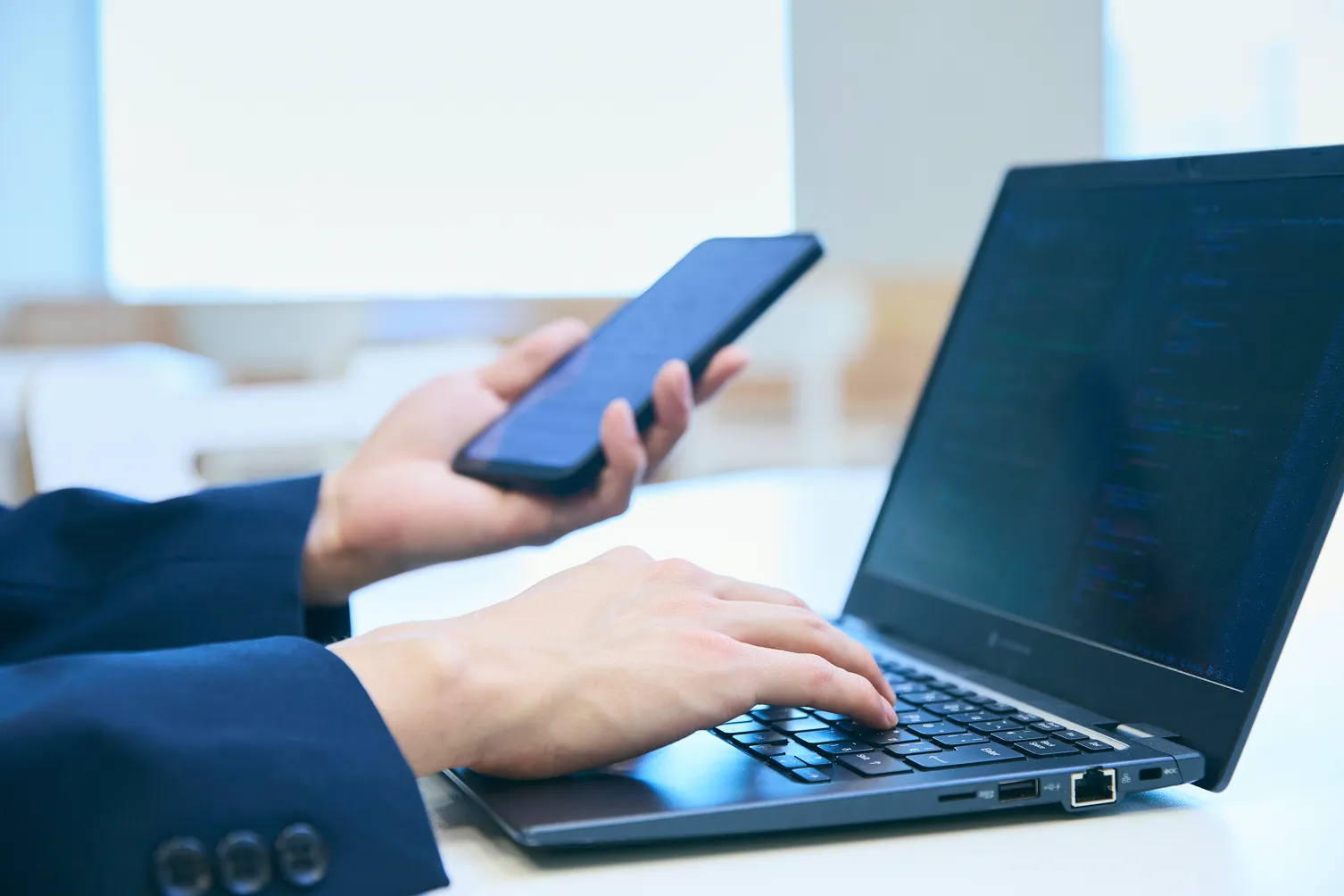 チーム統括 T.K.さんがデスクでスマートフォンを片手に持ちPCを操作している写真