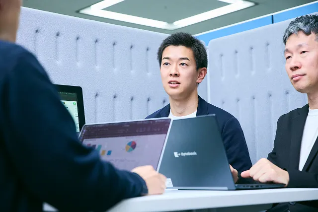 若手男性社員3人が、ホワイトボードを見ながら打ち合わせをしている写真。