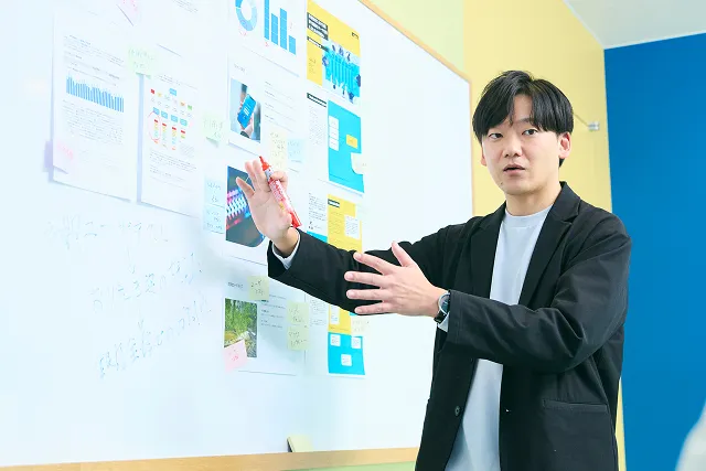 若手男性社員が、ホワイトボードの前でプレゼンをしている写真。
